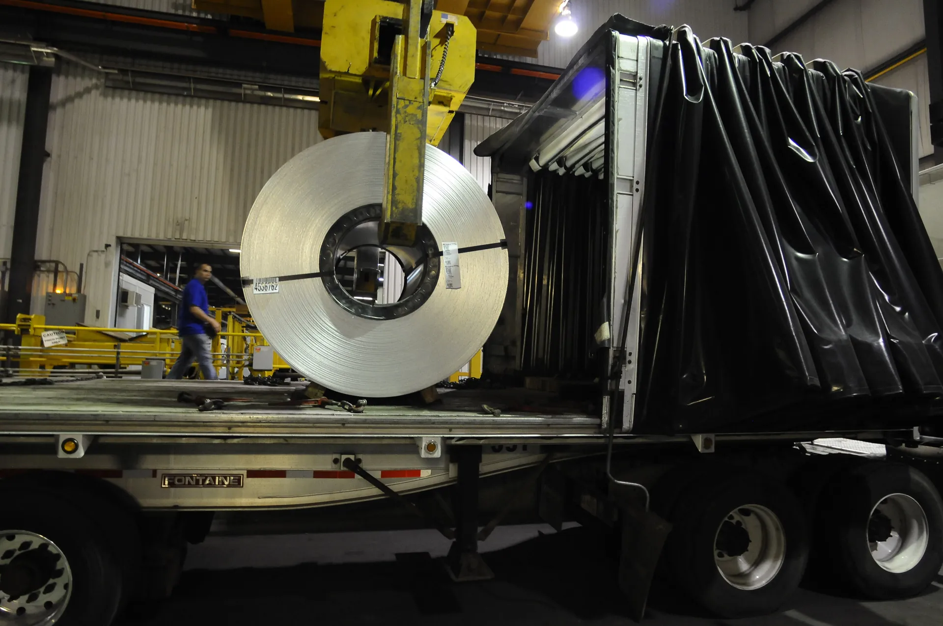 Crane C-hook lowering a steel coil onto a flatbed truck inside a service center
